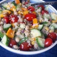 A bowl of colorful quinoa Greek salad with chickpeas, cherry tomatoes, cucumbers, bell peppers, and corn sits with a spoon on a dark blue tablecloth.