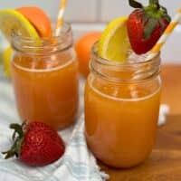 Two mason jars filled with orange juice, each garnished with a lemon slice and a strawberry, with striped straws. A fresh strawberry and orange are nearby, on a wooden surface with a striped cloth.