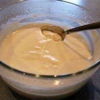A clear glass bowl filled with creamy white homemade yogurt, with a metal spoon resting inside, placed on a gray countertop.