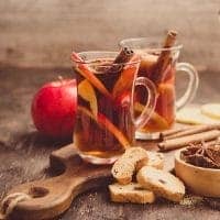 Two glass mugs of mulled wine with apple slices and cinnamon sticks sit on a wooden board with cookies, spiced tea, star anise, and a red apple in the background.