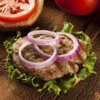 Open hamburger with a grilled beef patty, lettuce, and three rings of raw red onion on a whole wheat bun—like a classic turkey burgers recipe. Sliced tomato and top bun are beside it on a wooden surface.