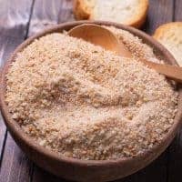 A wooden bowl filled with homemade breadcrumbs and a wooden spoon, with slices of bread in the background on a rustic wooden surface.