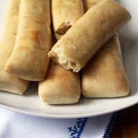 A white plate holds five golden-brown homemade breadsticks, one with a bite taken out. The plate rests on a white cloth with blue embroidery. The fivespot green living logo appears in the bottom right corner.