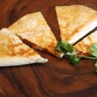 Four triangular slices of Mango and Brie Quesadilla are arranged on a wooden plate, garnished with fresh cilantro. The quesadillas appear golden brown and lightly toasted, with some visible filling. Fivespot Green Living logo is in the top left corner.