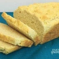 A loaf of gluten-free homemade bread rests on a teal cloth, partially sliced with three pieces fanned out in front. The bread has a golden crust and soft, crumbly texture. The Fivespot Green Living logo is in the bottom right corner.