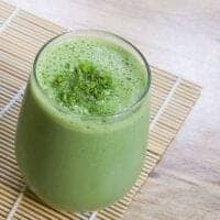 A glass of frothy green matcha green tea smoothie latte topped with matcha powder, placed on a bamboo mat and a light wooden surface.