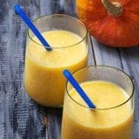 Two glasses filled with a creamy yellow pumpkin pie smoothie, each with a blue straw. The glasses rest on a rustic wooden surface, with a pumpkin partially visible in the background.