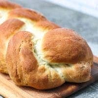 A golden-brown loaf of braided bread sits on a wooden cutting board, its soft, fluffy texture hinting at a Cardamom Homemade Bread Recipe. The fresh, homemade loaf rests on a gray countertop, emitting an inviting aroma.