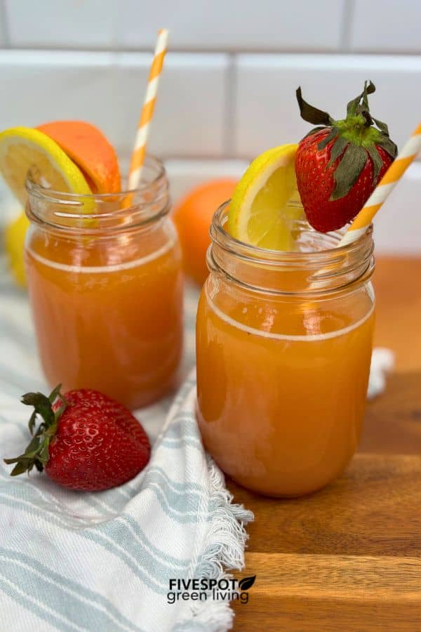 Two mason jars filled with orange juice, each garnished with a lemon slice and a strawberry, with striped straws. A fresh strawberry and orange are nearby, on a wooden surface with a striped cloth.